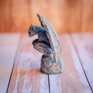 May include: A gray gargoyle statue with large wings, perched on a stone base. The statue has a detailed face and is in a crouching position. The background is a blurred wooden surface.