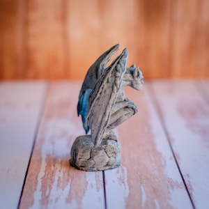 May include: A gray gargoyle statue with large, detailed wings and a crouching pose. The statue is made of a stone-like material and sits on a textured base. The wings have a hint of blue. The background is a light brown.