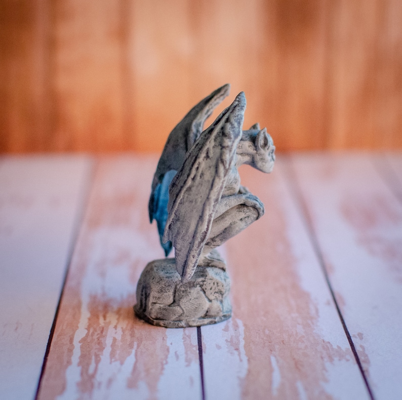 May include: A gray gargoyle statue with large, detailed wings and a crouching pose. The statue is made of a stone-like material and sits on a textured base. The wings have a hint of blue. The background is a light brown.