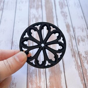 May include: Black circular cutout with a decorative design resembling a stained glass window. The design features a central circle with six pointed arches radiating outwards.