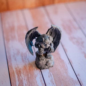 May include: A small, gray gargoyle statue with outstretched wings. The figurine is perched on a stone base, with detailed features and a weathered appearance. The background is a light wood surface.
