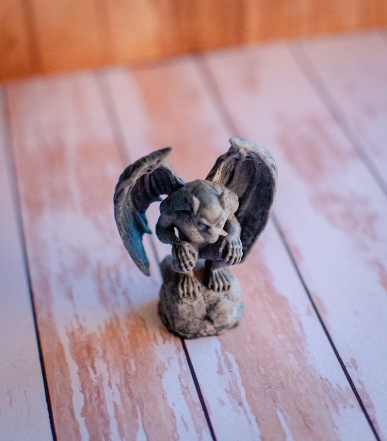 May include: A small, gray gargoyle statue with outstretched wings. The figurine is perched on a stone base, with detailed features and a weathered appearance. The background is a light wood surface.