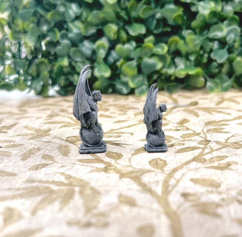 Może przedstawiać: Dwie małe, szare posągi gargulc&oacute;w. Każdy posąg przedstawia skrzydlatą istotę siedzącą na globie, o szczeg&oacute;łowym, teksturowanym wyglądzie. Posągi są ustawione na tle zielonych liści i wzorzystej powierzchni.
