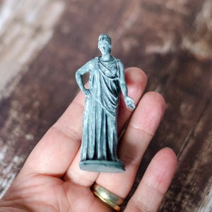 K&ouml;nnte beinhalten: Eine kleine, graue Statue einer bekleideten Figur, m&ouml;glicherweise eine klassische G&ouml;ttin, mit Augenbinde. Die Statue wird in einer Hand gehalten und zeigt ihre komplizierten Details und ihr Design. Die Figur hat einen Arm an der Seite und den anderen am Ellbogen angewinkelt.