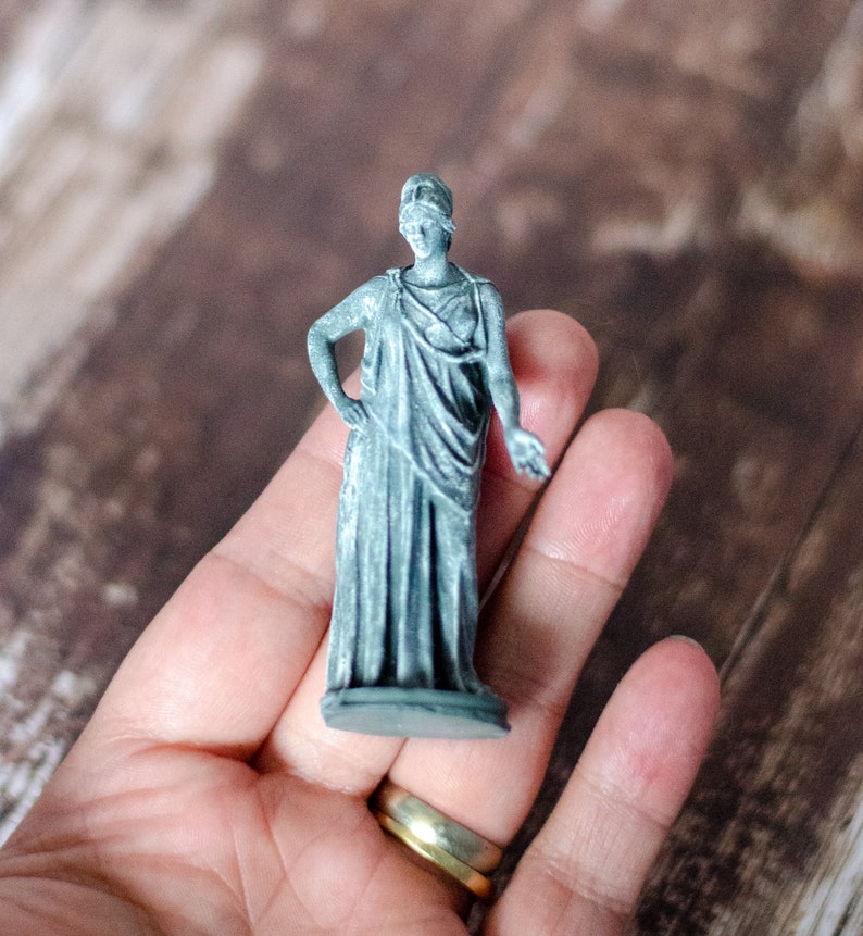 K&ouml;nnte beinhalten: Eine kleine, graue Statue einer bekleideten Figur, m&ouml;glicherweise eine klassische G&ouml;ttin, mit Augenbinde. Die Statue wird in einer Hand gehalten und zeigt ihre komplizierten Details und ihr Design. Die Figur hat einen Arm an der Seite und den anderen am Ellbogen angewinkelt.