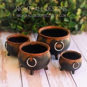 May include: A set of four black and rust-colored miniature cauldrons with three legs and a silver ring handle on each side. The text NIGHTFALL DESIGNS and NIGHTFALLDESIGNSCO.ETSY.COM is visible at the top of the image above the cauldrons.
