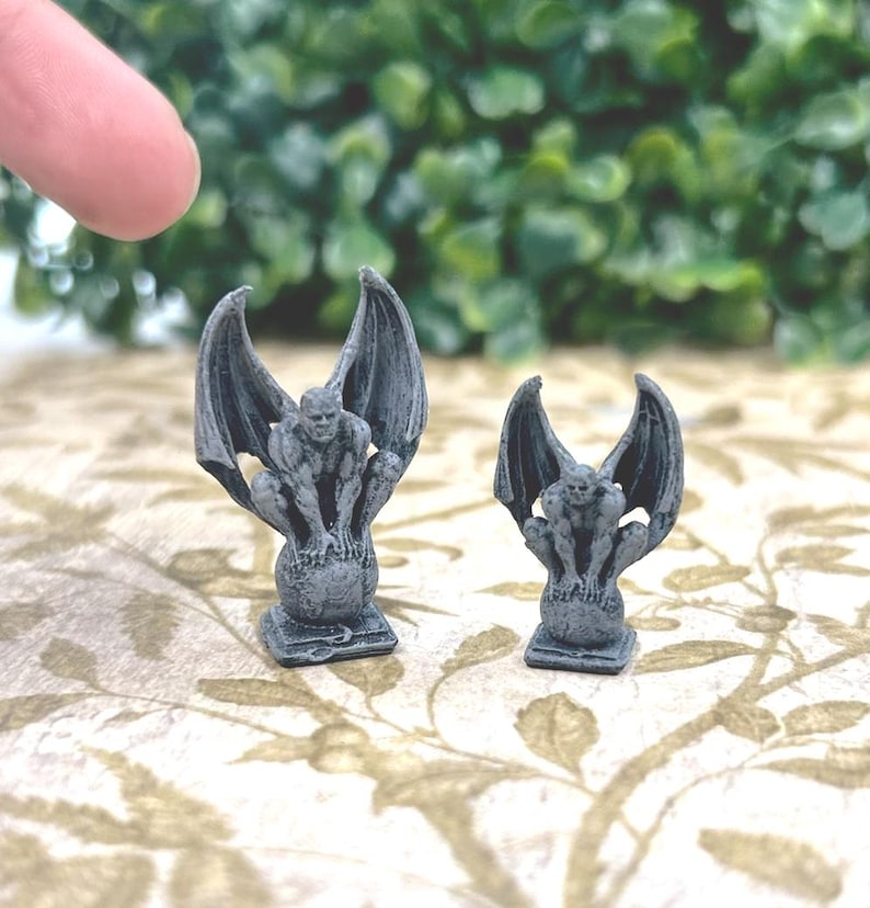 May include: Two gray gargoyle figurines with outstretched wings. The gargoyles are perched on a globe-like base. The figurines are detailed with a textured appearance, and are displayed on a patterned surface.
