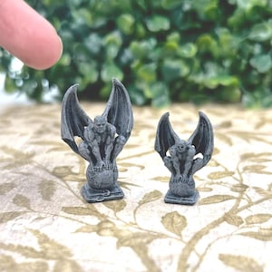 May include: Two gray gargoyle figurines with outstretched wings. The gargoyles are perched on a globe-like base. The figurines are detailed with a textured appearance, and are displayed on a patterned surface.