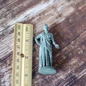 K&ouml;nnte beinhalten: Kleine, graue Statue einer bekleideten Figur, etwa 5 cm hoch, steht auf einem kreisf&ouml;rmigen Sockel. Die Figur posiert mit einem Arm an der Seite und dem anderen ausgestreckt. Ein Lineal mit Zollma&szlig;en befindet sich daneben.