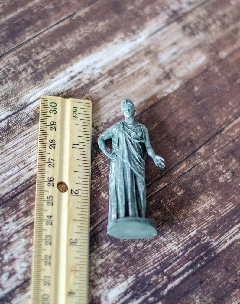 K&ouml;nnte beinhalten: Kleine, graue Statue einer bekleideten Figur, etwa 5 cm hoch, steht auf einem kreisf&ouml;rmigen Sockel. Die Figur posiert mit einem Arm an der Seite und dem anderen ausgestreckt. Ein Lineal mit Zollma&szlig;en befindet sich daneben.