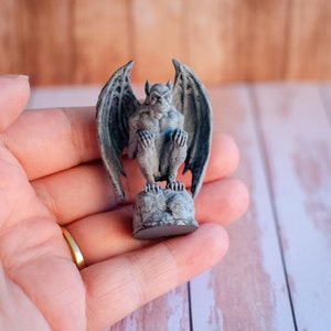 May include: A small, gray gargoyle statue with large, detailed wings, perched on a rocky base. The statue has a stern expression and is painted with subtle shading. The figurine is a decorative item, likely for home decor or a collector's piece.