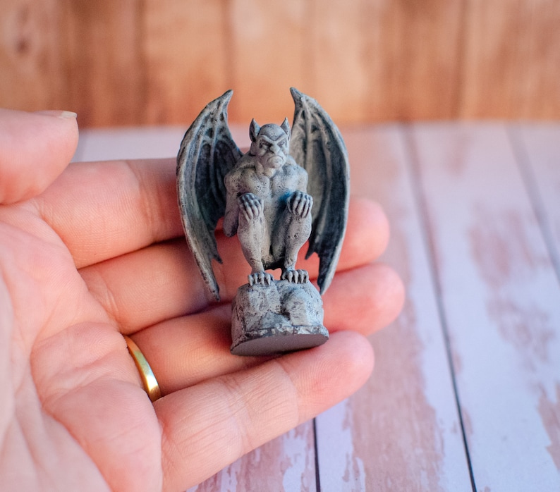 May include: A small, gray gargoyle statue with large, detailed wings, perched on a rocky base. The statue has a stern expression and is painted with subtle shading. The figurine is a decorative item, likely for home decor or a collector's piece.