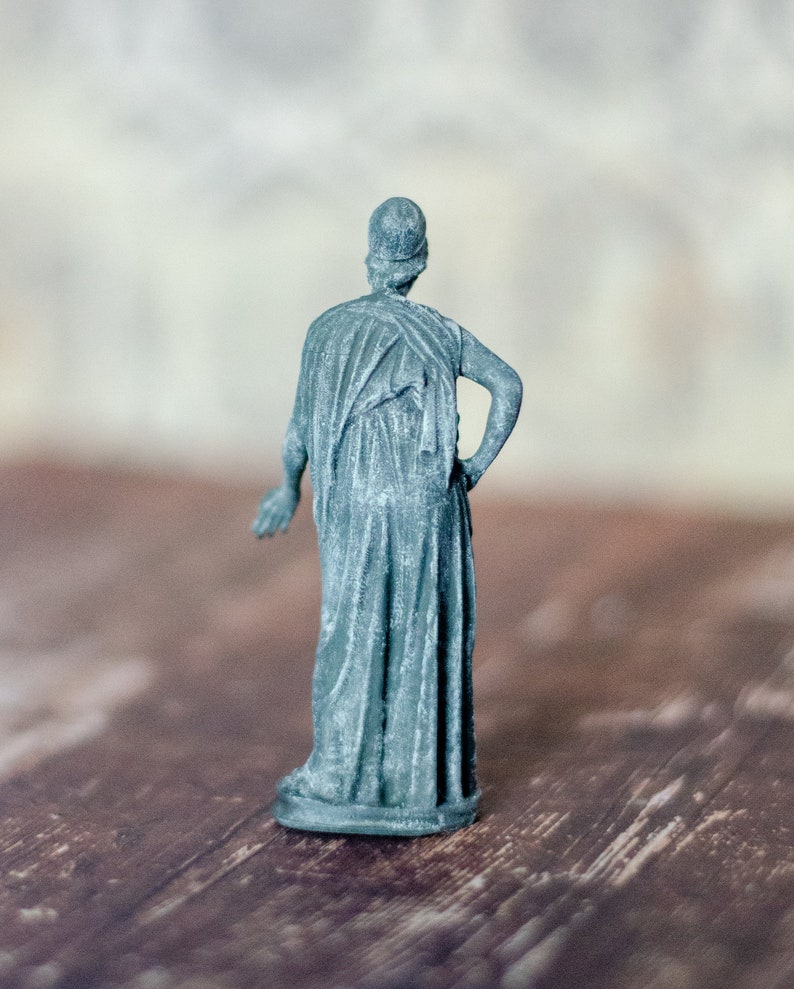 K&ouml;nnte beinhalten: Kleine, teal-farbene Statue einer klassischen Figur, von hinten gesehen. Die Figur ist in flie&szlig;ende Gew&auml;nder geh&uuml;llt, ein Arm an der Seite, der andere in der H&uuml;fte. Die Statue steht auf einer Holzoberfl&auml;che.