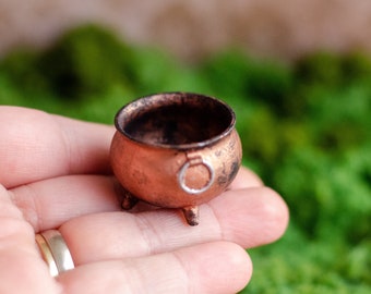 May include: A small, copper-colored cauldron with three short legs and a small handle. The cauldron has a dark interior and a slightly tarnished finish, suggesting an aged appearance. The miniature pot is likely used for decorative or crafting purposes.