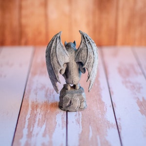 May include: A gray gargoyle statue with large, detailed wings, perched on a stone base. The statue has a textured surface and is viewed from the back. The background is a blurred wooden surface.