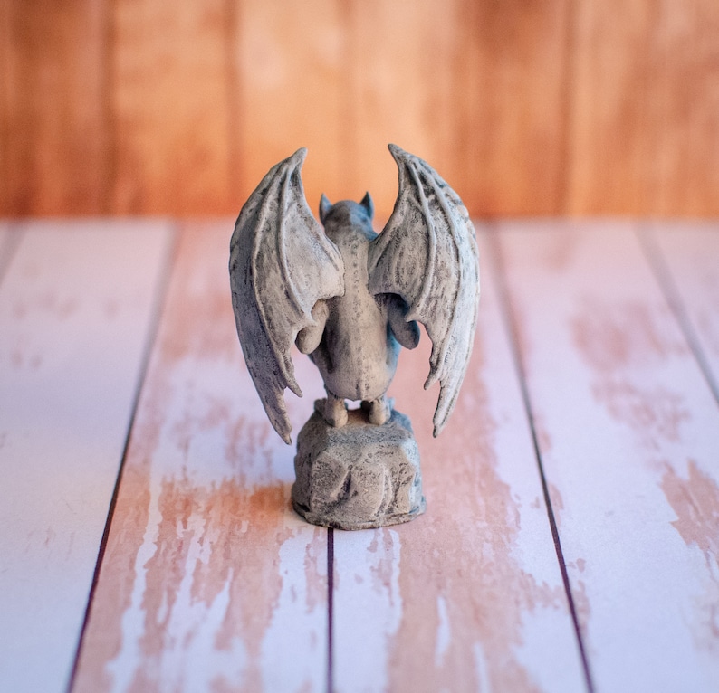 May include: A gray gargoyle statue with large, detailed wings, perched on a stone base. The statue has a textured surface and is viewed from the back. The background is a blurred wooden surface.