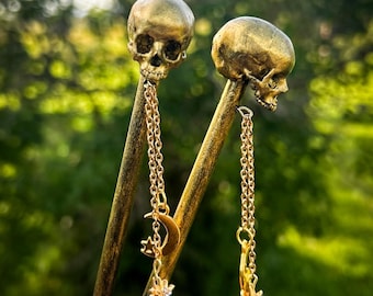 May include: Two golden hair sticks, each topped with a skull and dangling chain charms. One chain features a crescent moon, star, and a flower-like charm. The other has a star-shaped charm.