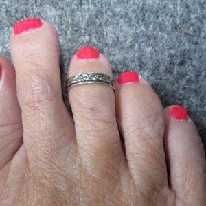 May include: Close-up of a foot with red nail polish on the toes. A silver toe ring with a detailed pattern is on the second toe. The skin tone is fair, and the background is a textured gray surface.