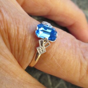 May include: A gold ring with a blue gemstone and white accents. The ring is on a finger.