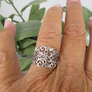 May include: A silver-colored ring with an openwork design featuring swirling patterns and leaf-like shapes. The ring is wide and covers a significant portion of the finger. The ring is shown on a hand with a neutral background.