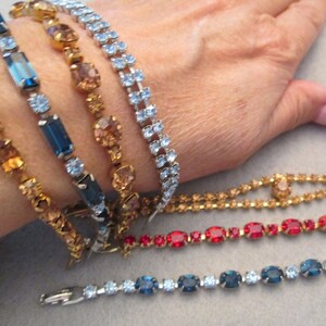 May include: Five rhinestone bracelets in various colors, including gold, blue, and red. The bracelets are stacked on a person's wrist.