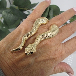 May include: A pair of gold snake rings. The rings are designed to wrap around two fingers. The snakes are detailed with scales and have black eyes.