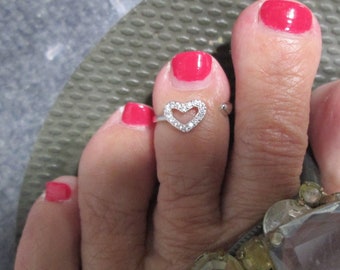 May include: A silver toe ring with a heart-shaped design featuring small clear rhinestones. The ring is worn on a toe with pink nail polish.