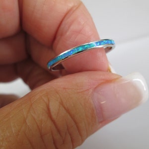 May include: A silver ring with a band of blue and green opal. The ring is held between fingers, showcasing the vibrant colors of the opal. The ring is a delicate piece of jewelry.