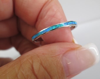 May include: A silver ring featuring a band of blue and green opal. The ring is held between fingers, highlighting the opal's colours. This delicate ring is a piece of jewellery.