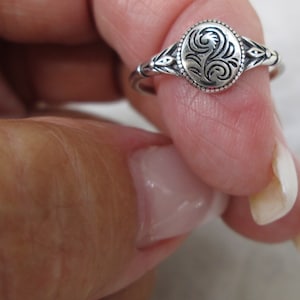 May include: A silver ring with an oval, engraved centerpiece featuring a swirling floral design. The ring has a detailed band with decorative elements on the sides. The ring is shown on a finger.