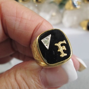 May include: A gold ring with a black onyx stone set in a square shape. The stone has a small diamond in the top corner and the letter "F" in gold lettering.