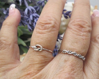 May include: Two silver rings on a hand. One ring is a simple knot design, the other is a twisted rope design.