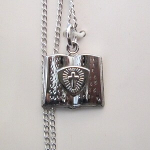 May include: Sterling silver necklace with a small open book pendant featuring a cross on a shield. The pendant is detailed with a textured surface.