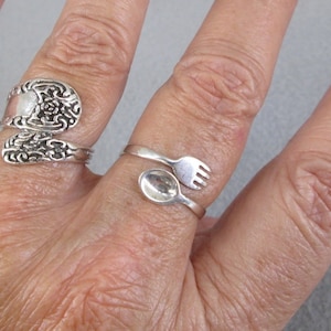 May include: Two silver rings, one with a spoon and fork design and the other with an ornate floral design, worn on a person's finger.