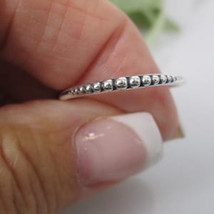 Peut inclure: Une bague en argent avec un motif de perles. La bague est fine et présente une série de petites perles rondes le long de l'anneau. La bague est tenue entre les doigts, mettant en valeur ses détails délicats. Un bijou simple et élégant.