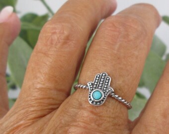 May include: A silver ring featuring a turquoise hamsa hand charm. The ring is worn on a finger.