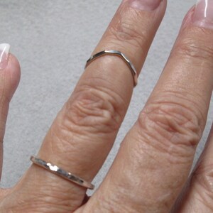 May include: A close-up of a hand wearing two silver rings. The ring on the index finger is a simple band, while the ring on the middle finger is a more intricate design.