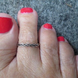 May include: Close-up of a foot with red nail polish and a silver braided toe ring. The foot is resting on a gray surface. The big toe and other toes are visible.