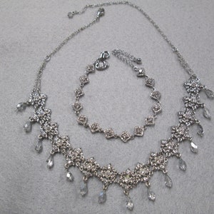 May include: A silver necklace and bracelet set with clear crystal accents. The necklace features a delicate chain with a floral design and dangling crystals. The bracelet is a simple chain with square-shaped crystals.