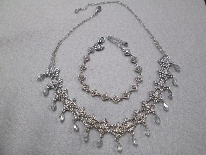 May include: A silver necklace and bracelet set with clear crystal accents. The necklace features a delicate chain with a floral design and dangling crystals. The bracelet is a simple chain with square-shaped crystals.