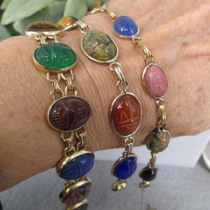 May include: Gold-toned bracelets with oval-shaped gemstone charms in various colors, including green, red, blue, and pink. The charms are connected by a delicate chain. The bracelets are displayed on a wrist.