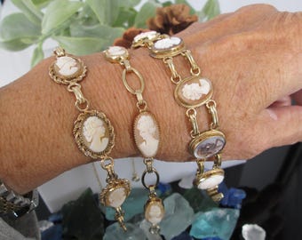 May include: Three gold-toned cameo bracelets with oval and round pendants are displayed. The cameos feature white relief portraits. The bracelets are connected by gold links. The bracelets are on a person's wrist.