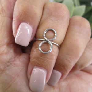 May include: A silver infinity symbol ring on a finger with light pink nail polish.