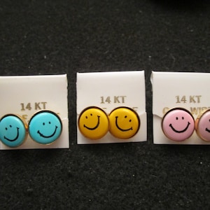 May include: Three pairs of 14KT gold smiley face earrings. The earrings are packaged on white cards with the text "14 KT" printed on them. The smiley faces are yellow, blue, and pink.