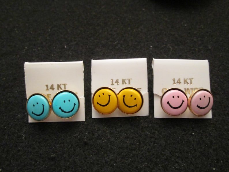 May include: Three pairs of 14KT gold smiley face earrings. The earrings are packaged on white cards with the text "14 KT" printed on them. The smiley faces are yellow, blue, and pink.