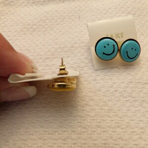 May include: A pair of gold-tone earrings with blue smiley face designs. The earrings are on a white card with the text "14KT" printed on it.