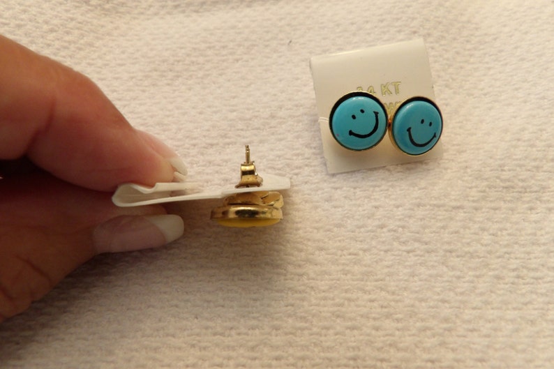 May include: A pair of gold-tone earrings with blue smiley face designs. The earrings are on a white card with the text "14KT" printed on it.