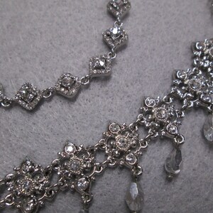 May include: Two silver necklaces with crystal accents. The top necklace has a simple design with square-shaped crystals. The bottom necklace has a more intricate design with a floral pattern and dangling crystals.
