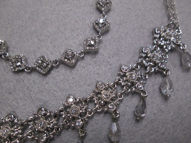 May include: Two silver necklaces with crystal accents. The top necklace has a simple design with square-shaped crystals. The bottom necklace has a more intricate design with a floral pattern and dangling crystals.