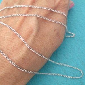 May include: A silver chain necklace with a curb link style. The chain is shown laid out on a person's arm.